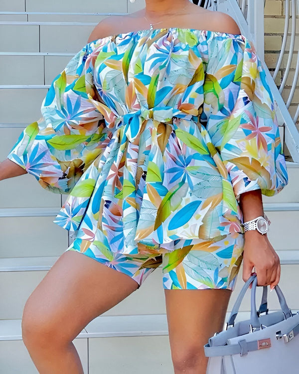 Une femme porte un ensemble short et haut à motifs wax. Le haut est à épaules dénudées et noué à la taille. Le short est taille haute et évasé. La femme est debout sur un perron et porte une montre au poignet gauche et un sac à main gris à la main droite.
