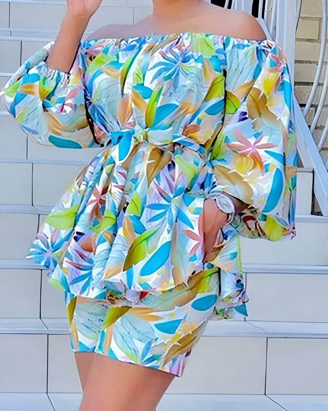 Une femme porte un ensemble short et haut à motifs wax. Le haut est à épaules dénudées et noué à la taille. Le short est taille haute et évasé. L'ensemble est de couleur bleu, jaune et rose.