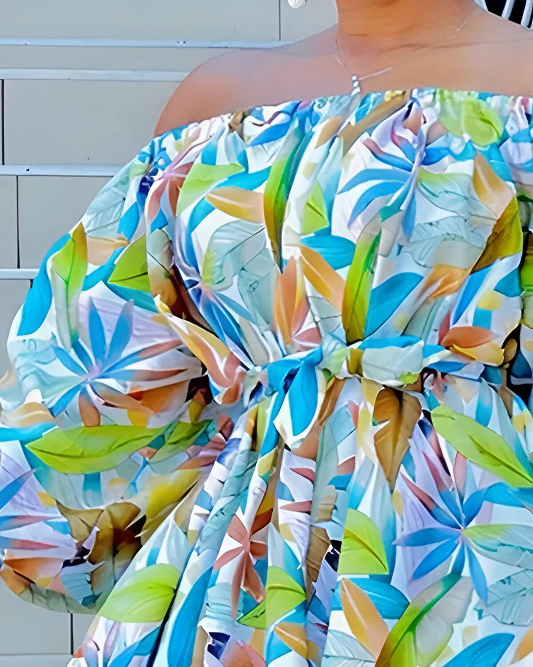 Une femme porte une robe colorée à motifs de feuilles tropicales. La robe est nouée à la taille et a des manches bouffantes. La femme est debout devant un mur blanc.