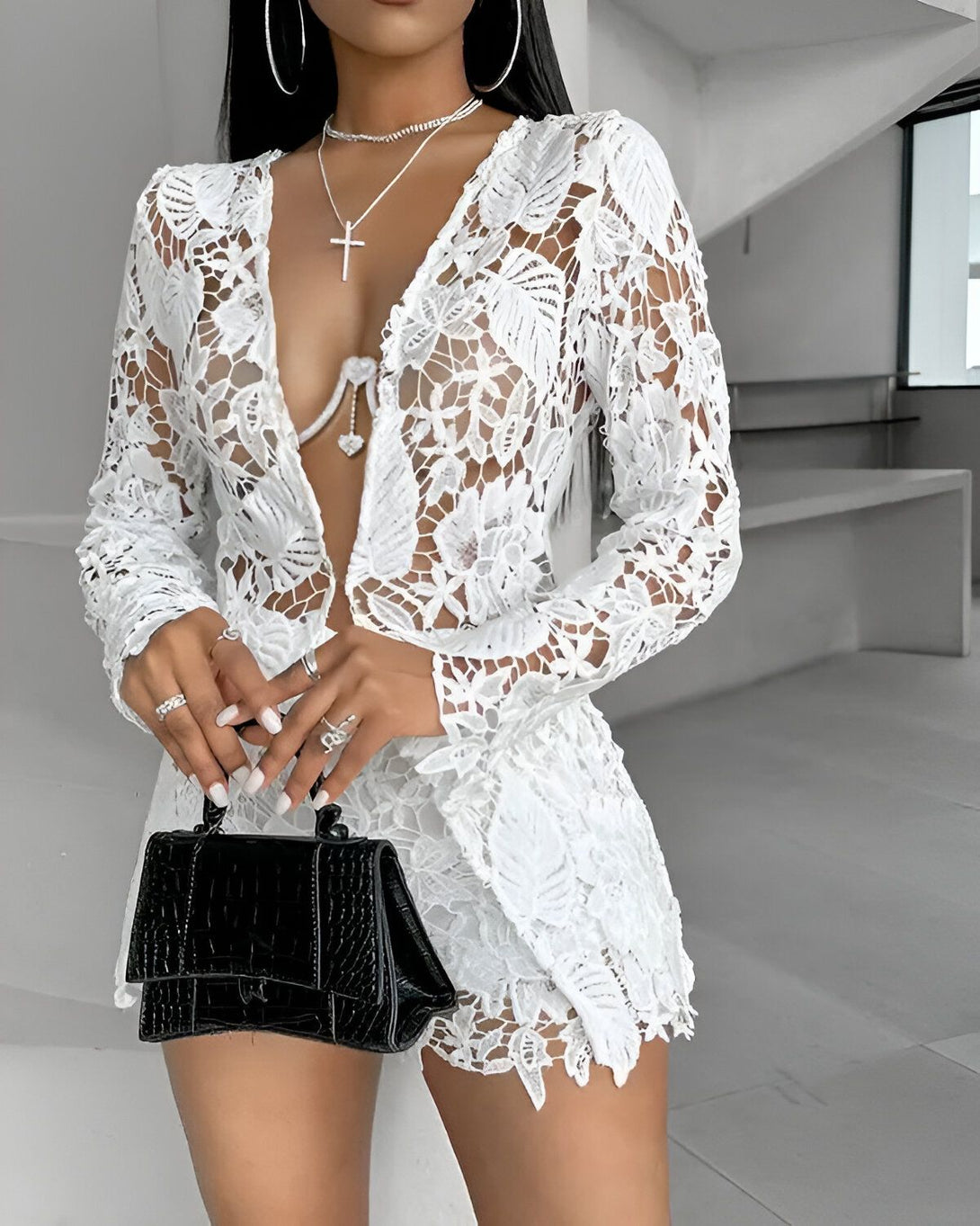 Une femme porte un ensemble de deux pièces en dentelle blanche. Le haut est un blazer avec un col en V profond et des manches longues. Le bas est un short taille haute avec une fermeture éclair sur le côté. La femme porte également un collier avec un pendentif en forme de croix, une bague à la main droite et un sac à main noir.