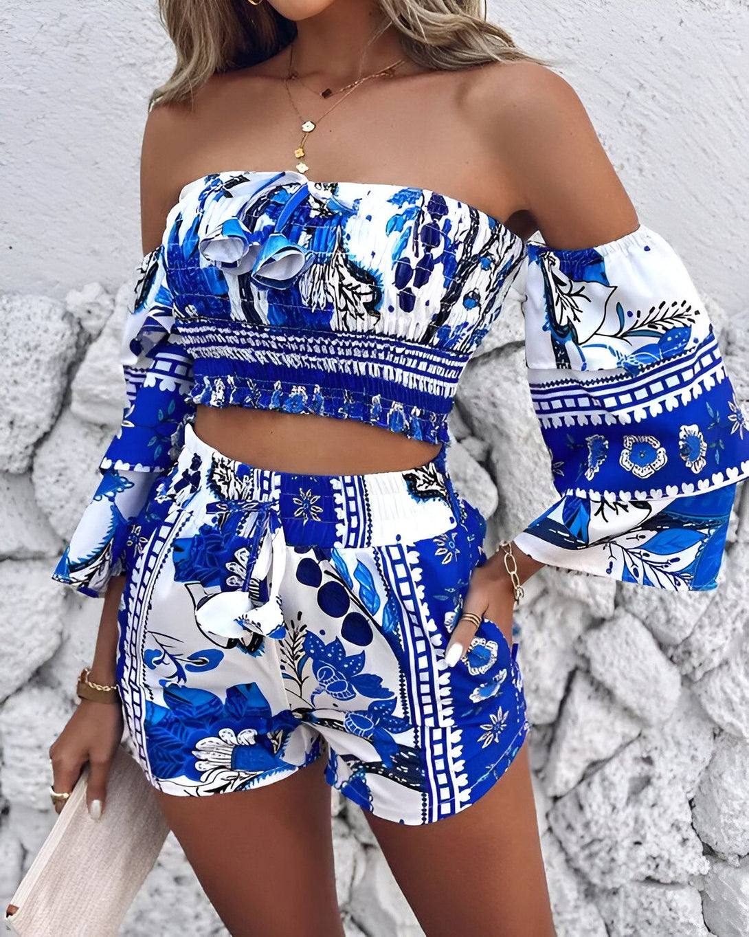 Une femme porte un ensemble deux pièces bleu et blanc. Le haut est un crop top à épaules dénudées avec des manches bouffantes. Le short est taille haute et possède des poches. L'ensemble est imprimé d'un motif floral.