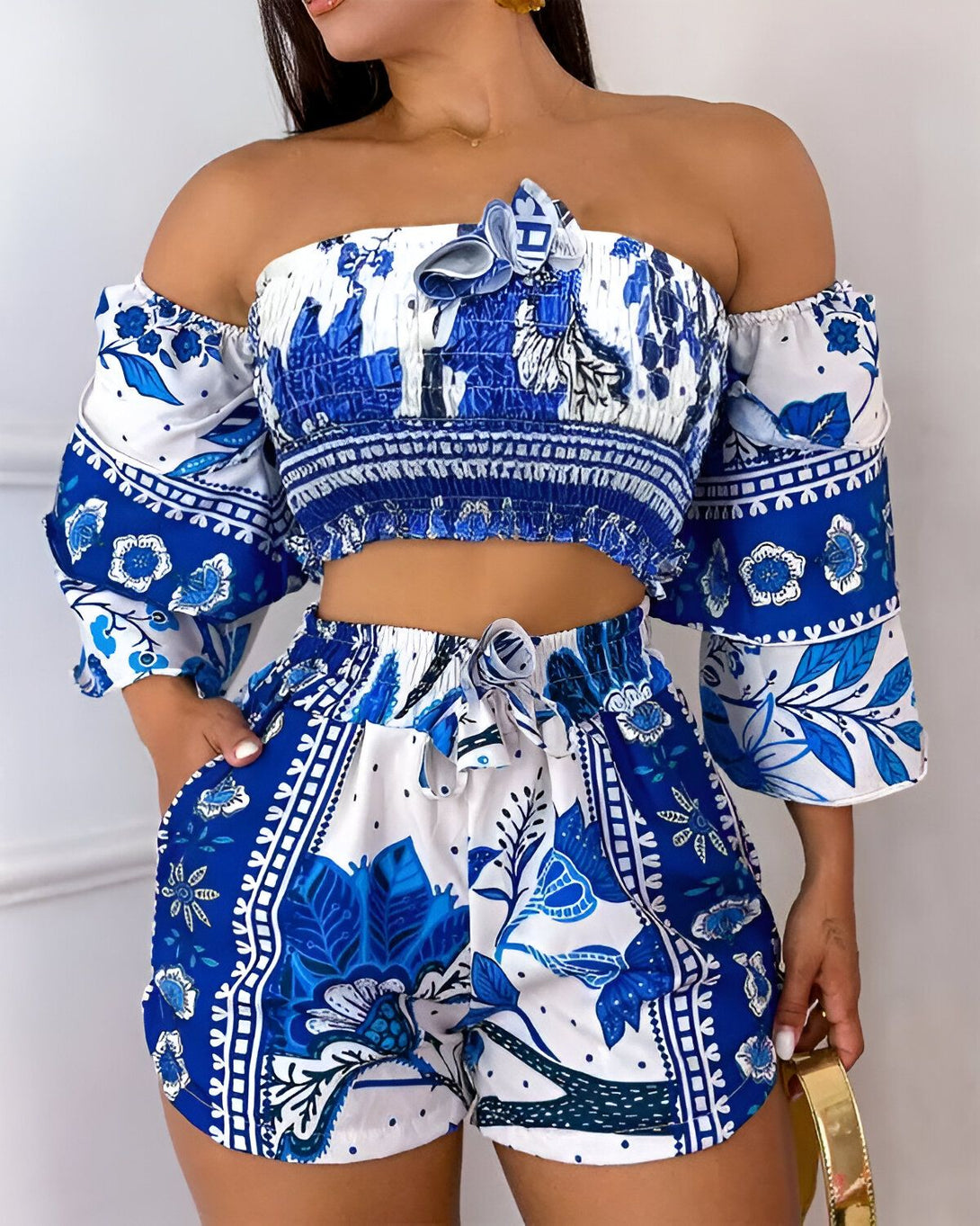 Une femme porte un ensemble deux pièces composé d'un crop top et d'un short. Le crop top est bleu et blanc, à épaules dénudées et à manches bouffantes. Le short est bleu et blanc, taille haute et à cordon de serrage. La femme est debout, les mains dans les poches du short. Elle porte également un sac à main doré.