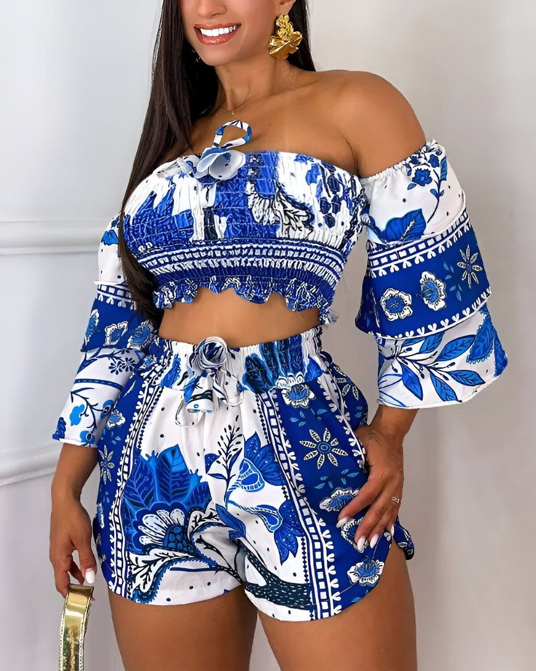Une femme porte un ensemble deux pièces bleu et blanc. Le haut est un crop top à épaules dénudées avec des manches bouffantes. Le short est taille haute et possède une ceinture à nouer. L'ensemble est imprimé d'un motif floral.