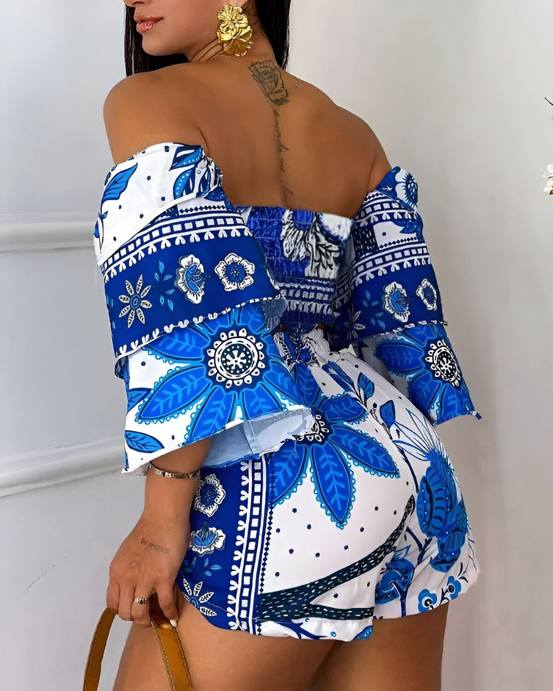 Une femme porte un ensemble deux pièces bleu et blanc. Le haut est à épaules dénudées et les manches sont bouffantes. Le short est taille haute et évasé. L'ensemble est imprimé de motifs floraux.
