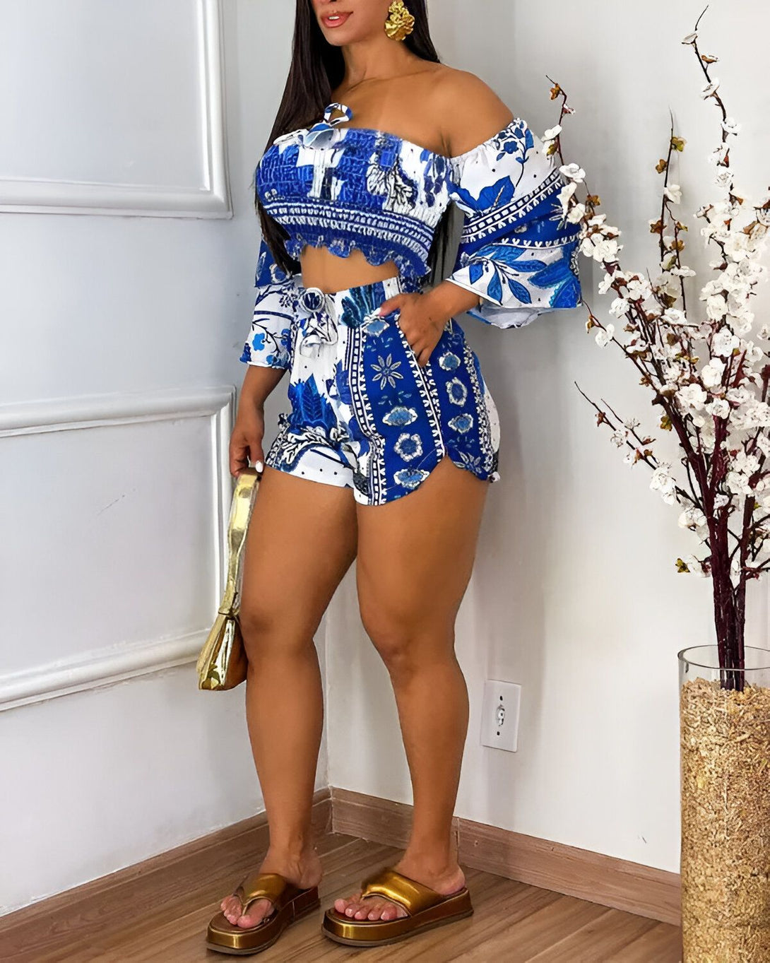 Une femme porte un ensemble short et haut court bleu et blanc. Le haut est à épaules dénudées et les manches sont bouffantes. Le short est taille haute et possède une ceinture. La femme est debout, les jambes légèrement écartées, et porte des sandales dorées. Elle a les cheveux longs et bruns et porte un maquillage léger.