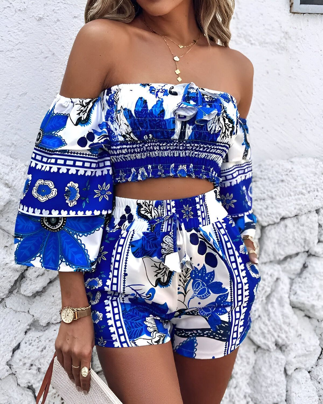 Une femme porte un ensemble deux pièces bleu et blanc. Le haut est un crop top à épaules dénudées et les manches sont bouffantes. Le short est taille haute et possède une ceinture à nouer.