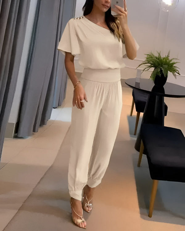 Une femme est debout dans un magasin. Elle porte un pantalon et un haut blancs. Le pantalon est ample et a une taille haute. Le haut a une manche courte et une épaule dénudée. La femme porte des sandales à talons hauts et un bracelet. Elle a les cheveux longs et bruns. Elle est en train de se prendre en photo dans un miroir.