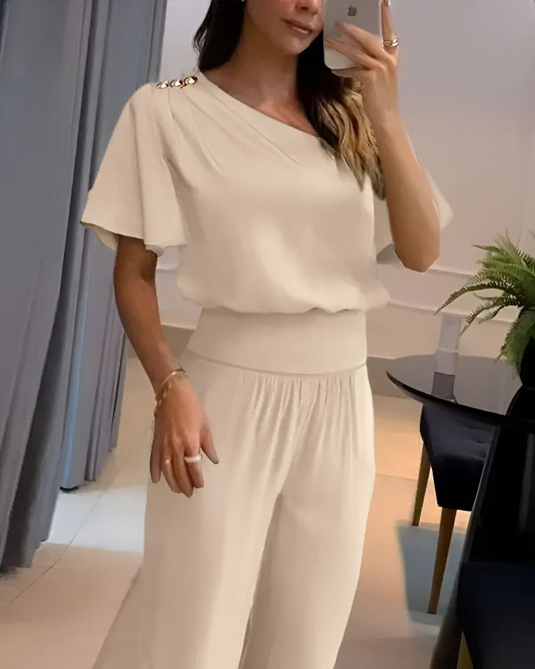 Une femme porte un pantalon et un chemisier blancs. Le chemisier a une manche courte et une épaule dénudée. Le pantalon est taille haute et évasé. La femme est debout dans une pièce, face à un miroir.