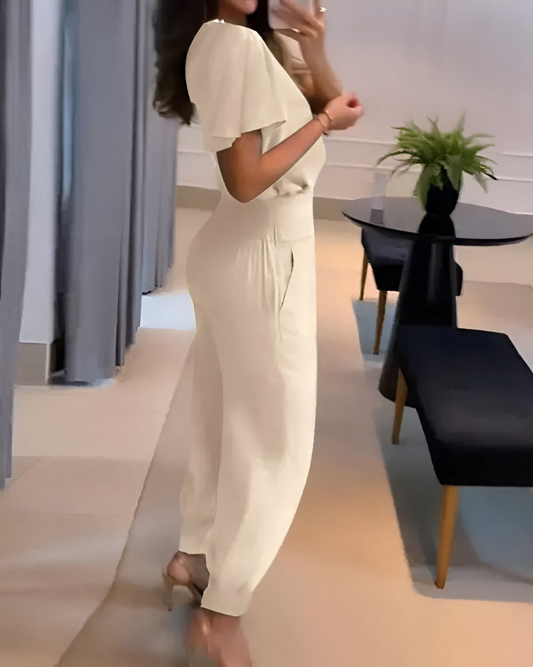 Une femme portant une combinaison pantalon beige à manches courtes et un décolleté en V. Elle est vue de dos et se trouve dans une pièce avec des rideaux gris et un sol en carrelage beige.