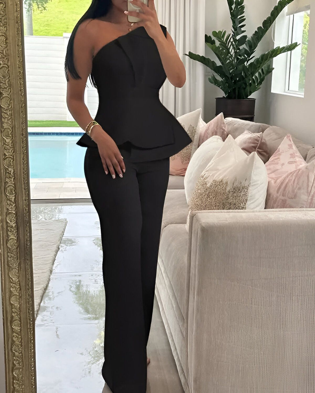 Une femme porte un ensemble pantalon et haut noir. Le haut est asymétrique et possède une manche. Le pantalon est large et taille haute.