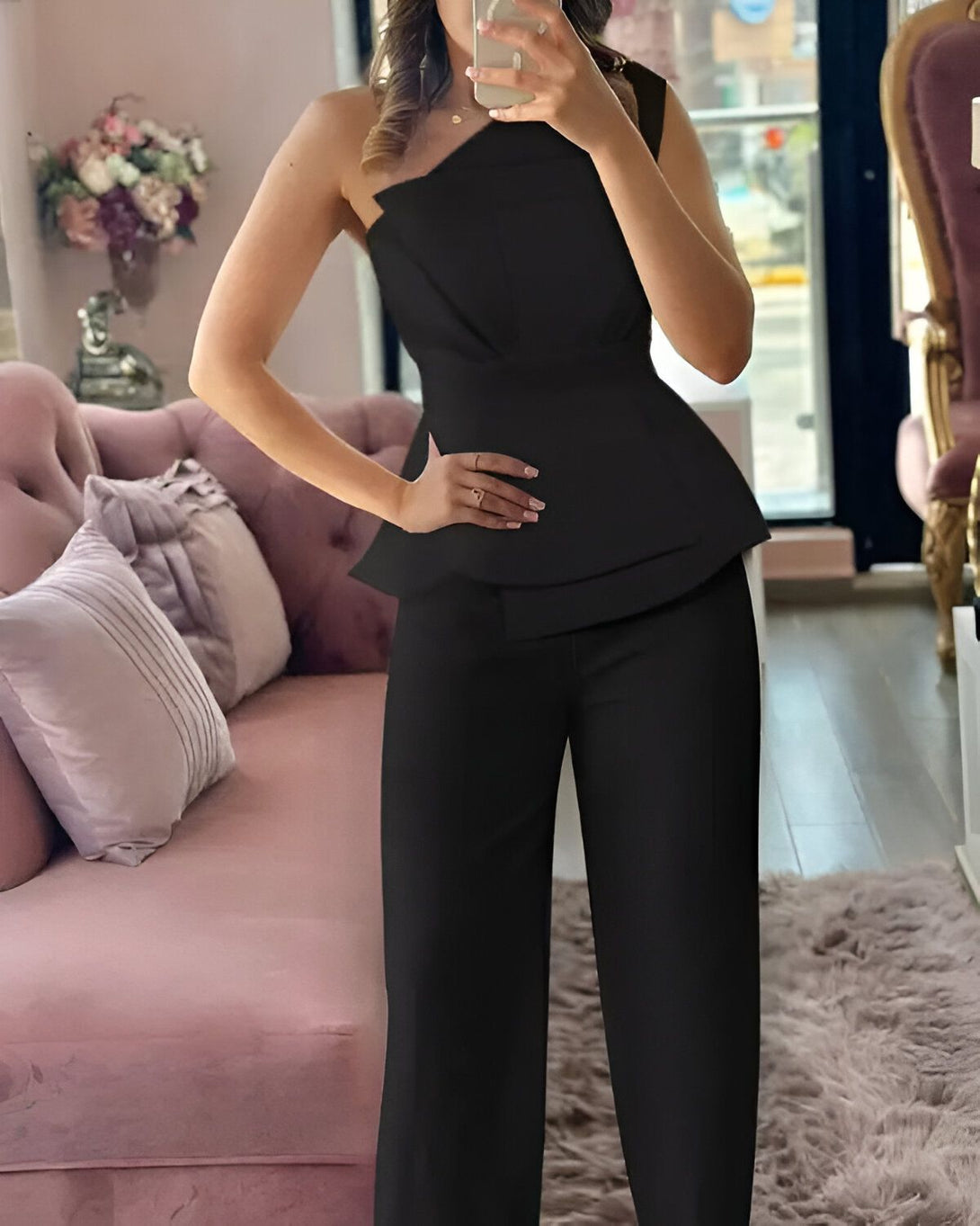 Une femme porte un tailleur pantalon noir. Le haut est asymétrique avec une manche et une épaule. Le pantalon est droit et taille haute.