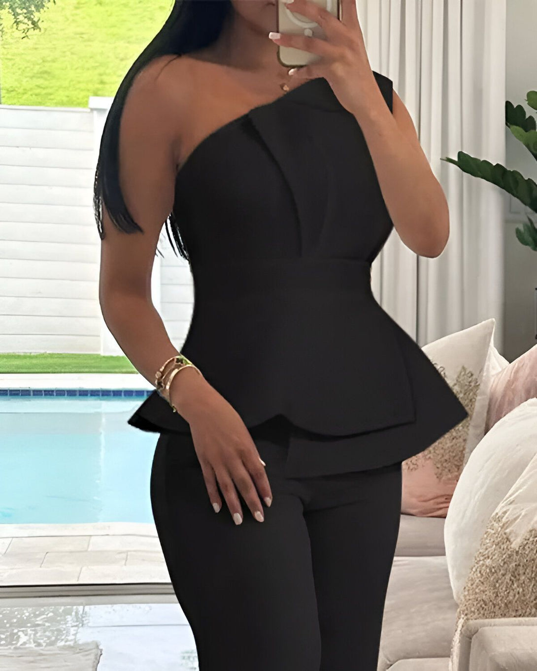 Une femme porte un ensemble pantalon et haut noir. Le haut est asymétrique avec une manche et une épaule. Le pantalon est moulant et taille haute.