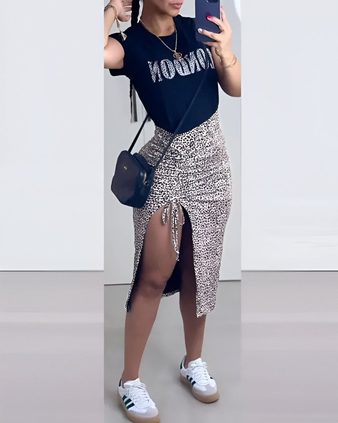 Une femme porte un t-shirt noir avec le texte "LONDON" en strass argentés, une jupe longue léopard fendue sur le côté droit, des baskets blanches et un sac à main noir.