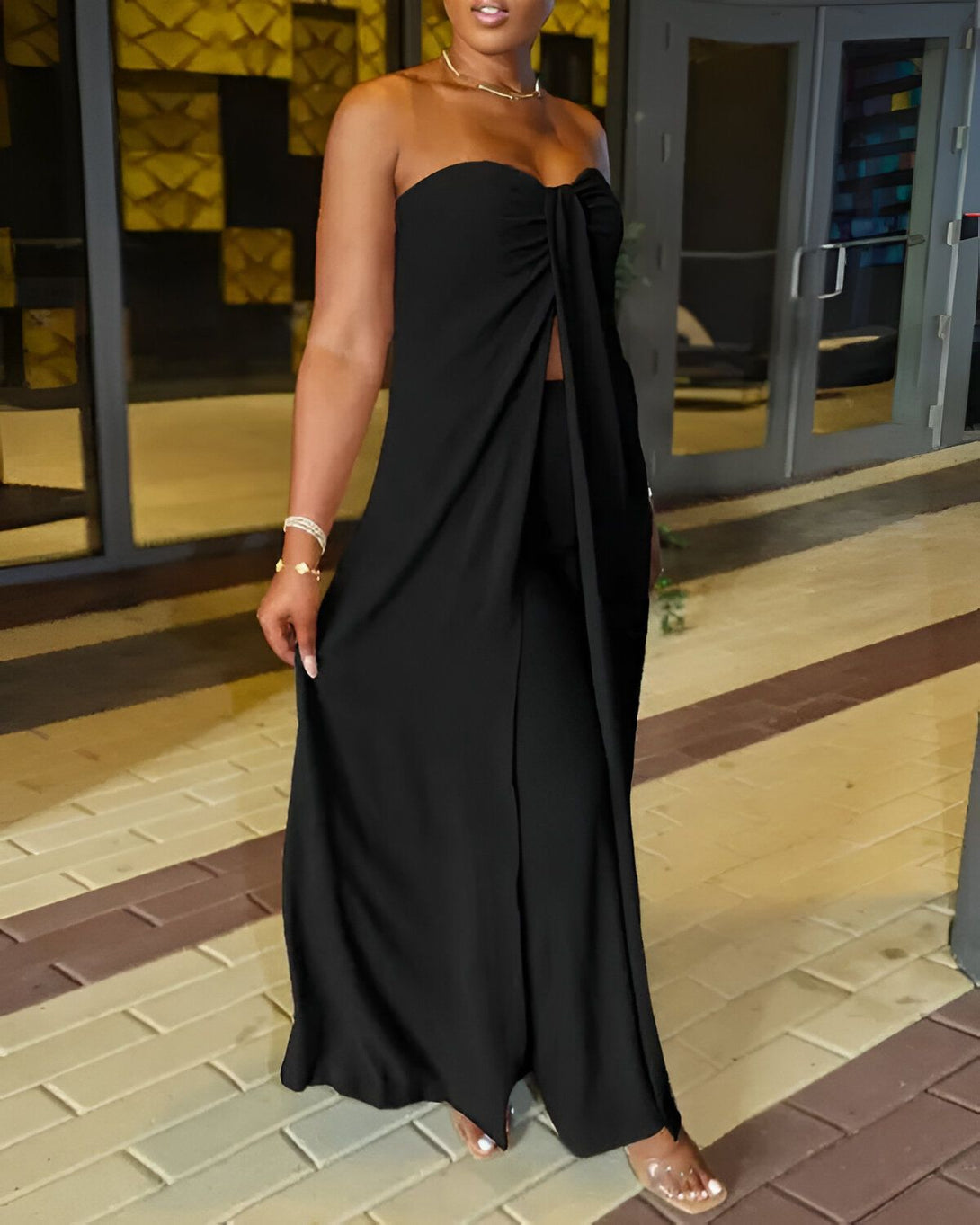 Une femme porte un ensemble pantalon large et haut bandeau noir. Elle est debout devant une porte vitrée.