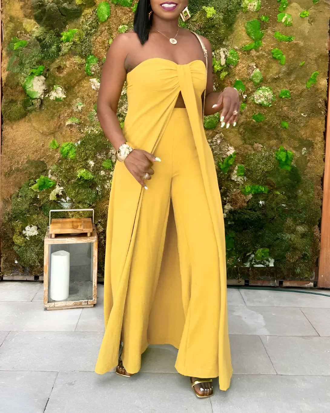 Une femme porte une combinaison pantalon jaune. Elle est debout devant un mur végétal.