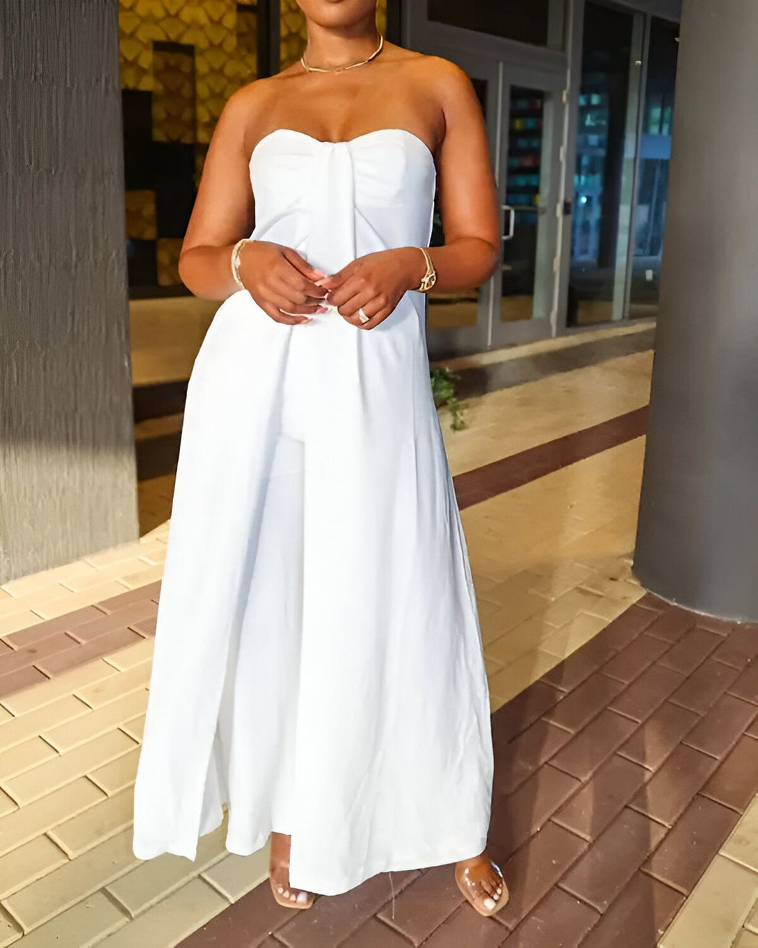 Une femme porte une combinaison pantalon blanche sans bretelles. Elle est debout dans un parking et porte des sandales marron clair.