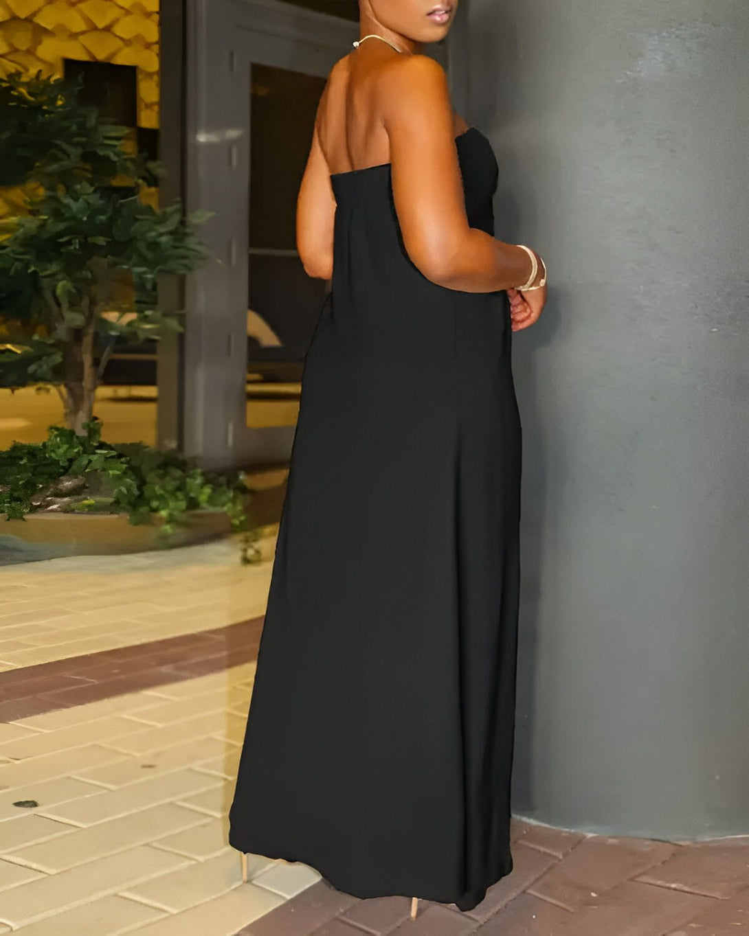 Une femme est debout, vue de dos. Elle porte une robe longue noire sans bretelles. Ses cheveux sont courts et noirs. Elle est pieds nus.