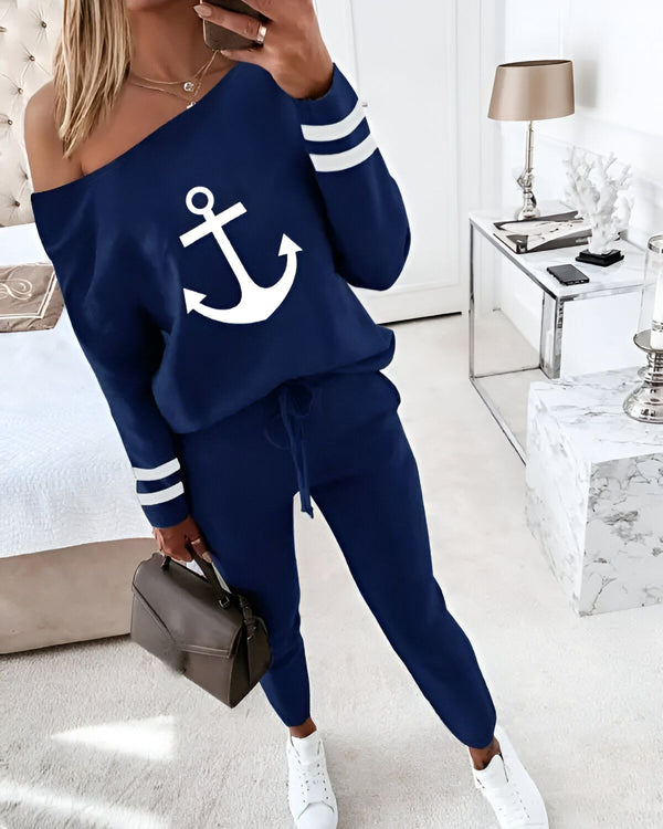 Une femme porte un ensemble de jogging bleu marine avec un motif d'ancre blanche sur le devant. Le haut est à épaules dénudées et les manches sont rayées blanches et bleues. Le pantalon est taille haute et possède une coulisse à la taille. La femme porte également des baskets blanches et un sac à main marron.