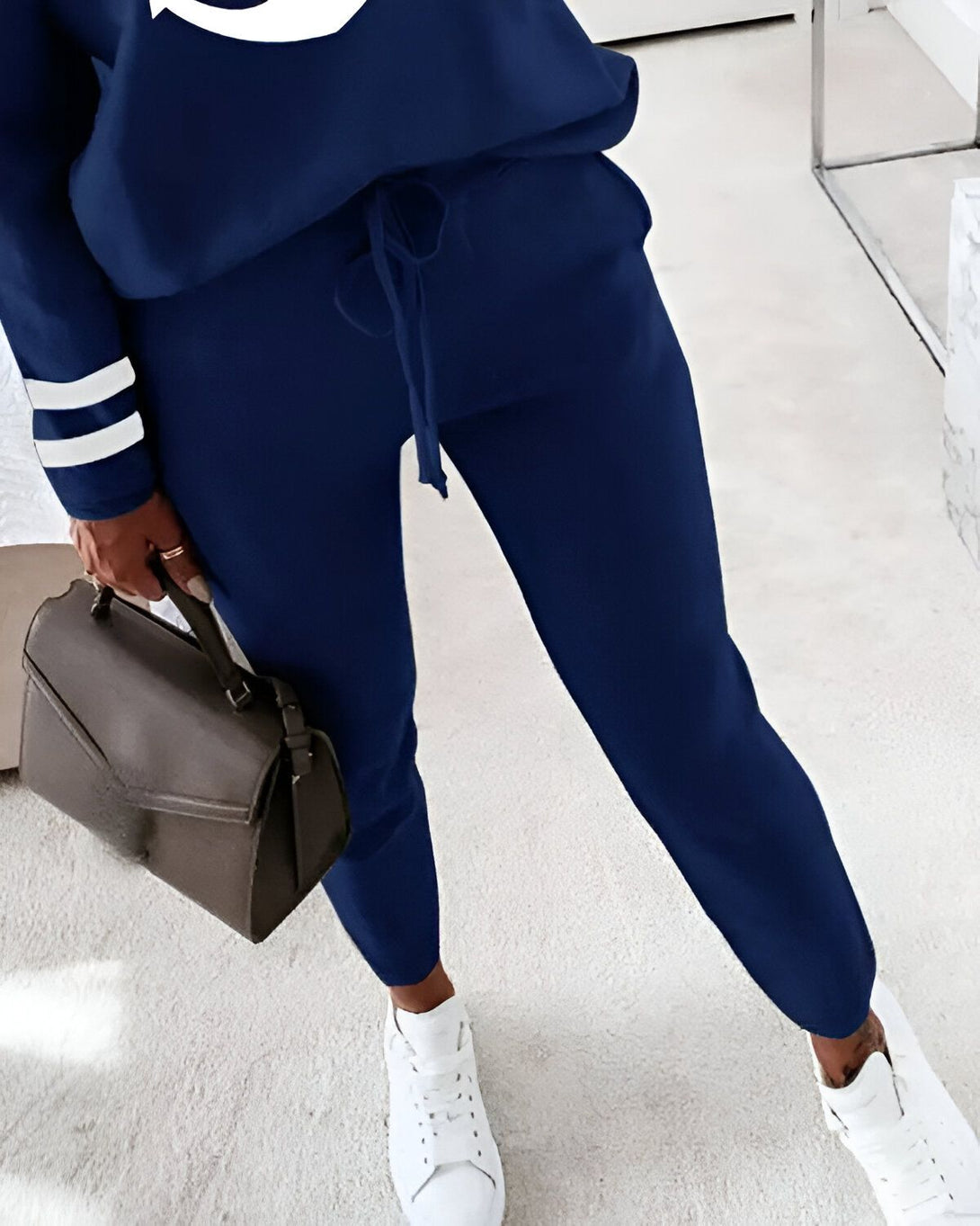 Une femme porte un survêtement bleu marine avec des baskets blanches et un sac à main marron.