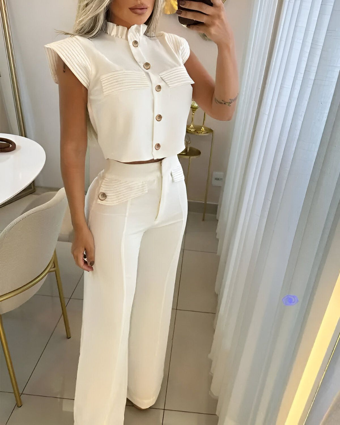 Une femme porte un chemisier blanc et un pantalon blanc. Le chemisier est court et a des manches bouffantes. Le pantalon est large et a une taille haute. La femme est debout dans une pièce et se prend en photo dans un miroir.