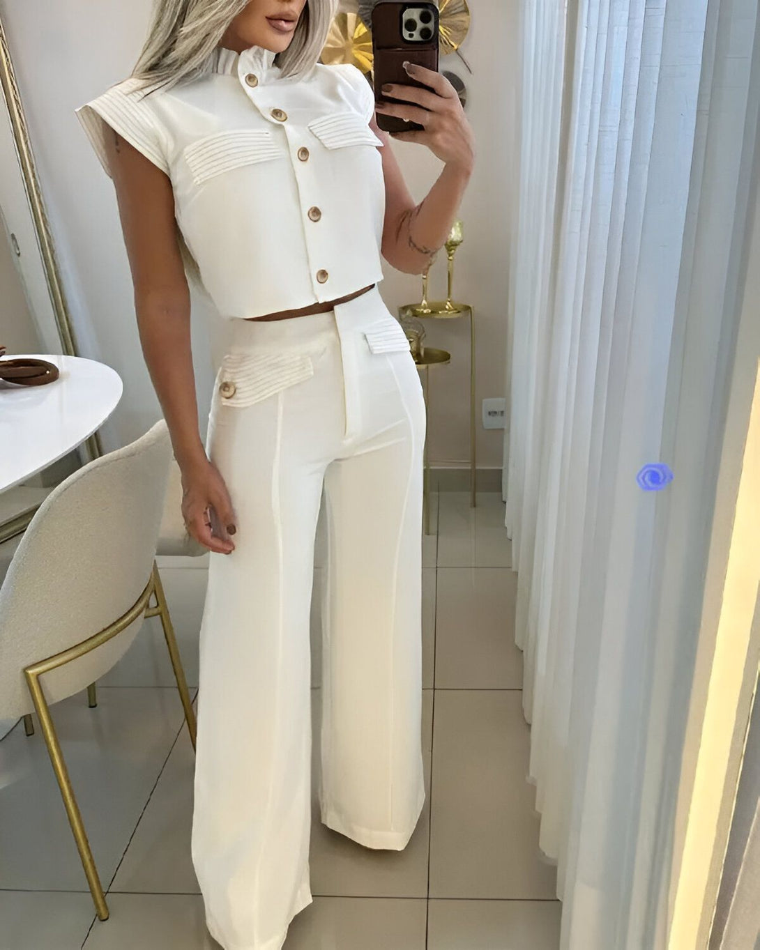 Une femme porte un chemisier blanc et un pantalon blanc. Le chemisier est court et a des manches bouffantes. Le pantalon est large et a une taille haute. La femme est blonde et a les cheveux longs. Elle est debout dans une pièce et se prend en photo avec son téléphone.