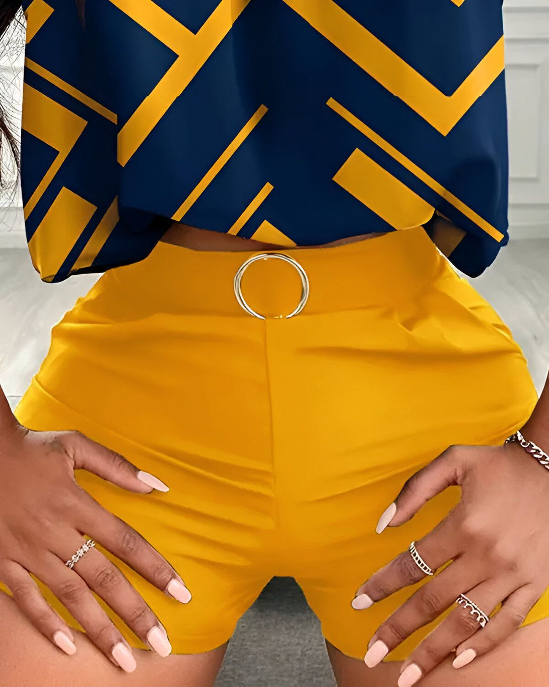 Une femme porte un ensemble deux pièces jaune et bleu. Le haut est court et sans manches, avec un motif géométrique. Le short est taille haute et possède une ceinture à anneau. La femme a les mains sur les hanches et porte des bagues aux doigts.