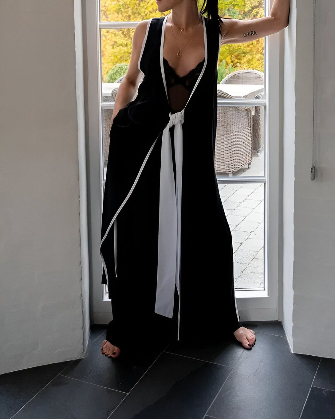 Une femme est debout dans l'embrasure d'une porte. Elle porte une combinaison pantalon noire et blanche. La combinaison est ouverte sur le devant et nouée à la taille. La femme est pieds nus.