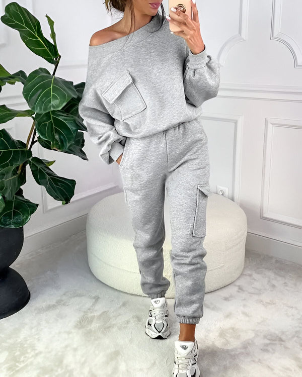 Une femme porte un survêtement gris avec une épaule dénudée et des poches. Elle porte également des baskets blanches.