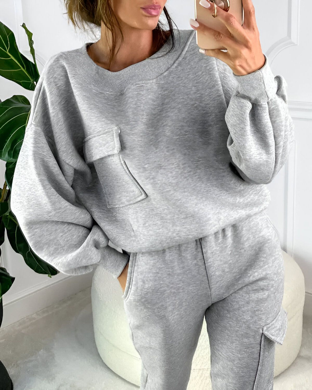 Une femme porte un survêtement gris composé d'un sweat-shirt à col rond et d'un pantalon de jogging. Le sweat-shirt a des poches et des manches bouffantes. Le pantalon a des poches cargo.
