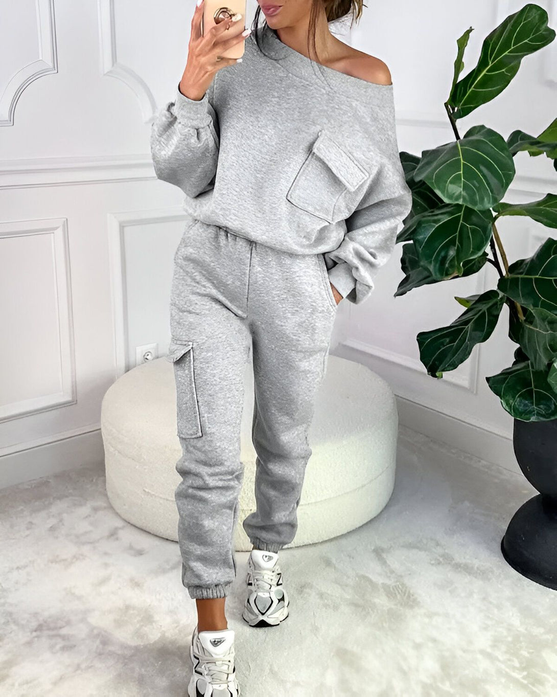 Une femme porte un survêtement gris avec une épaule dénudée et des poches sur le devant. Elle porte également des baskets blanches et se tient debout à côté d'une plante verte.