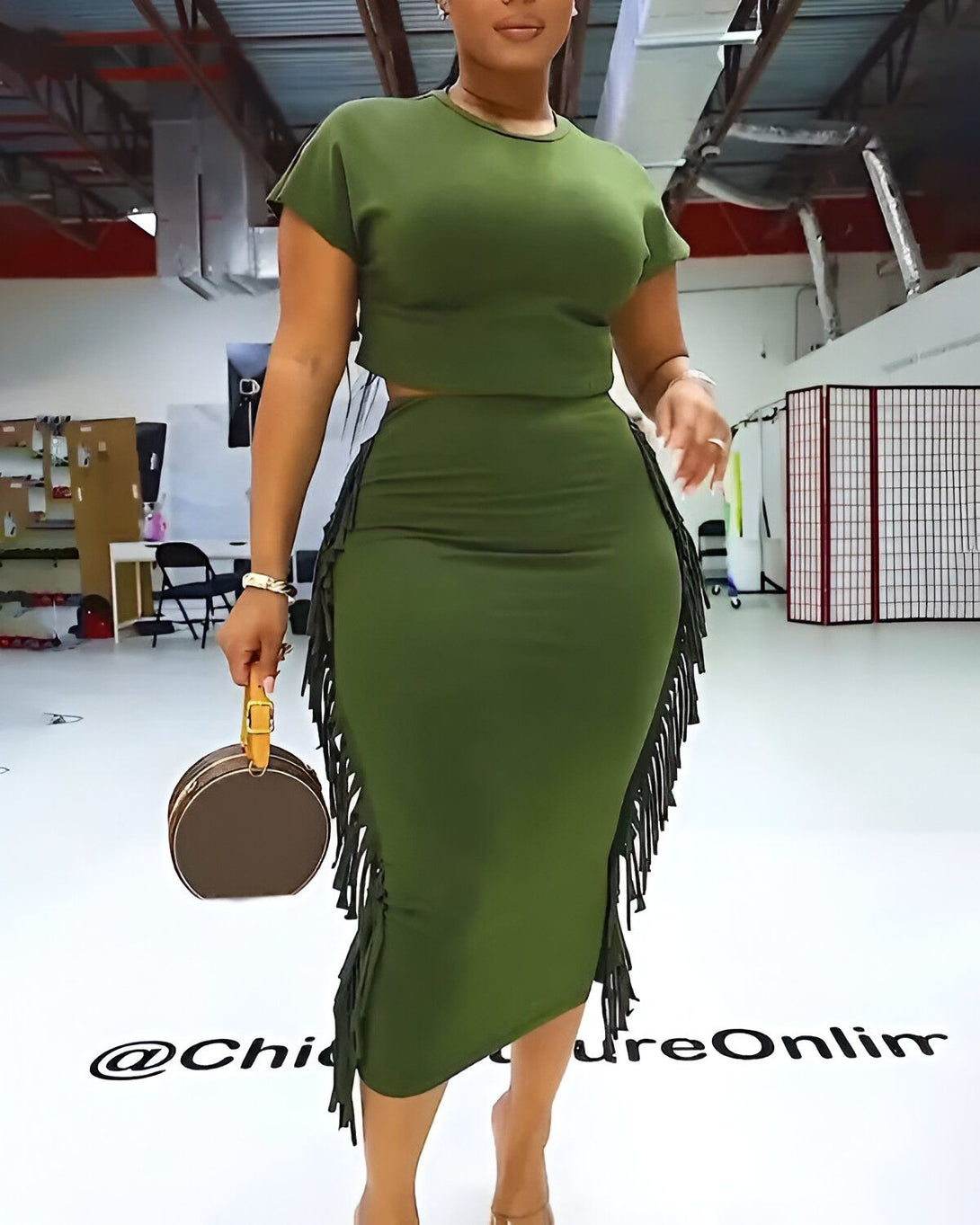 Une femme porte un ensemble deux pièces vert olive composé d'un crop top à manches courtes et d'une jupe crayon fendue sur le côté avec des franges. Elle porte également des sandales à talons couleur chair et un sac à main marron.