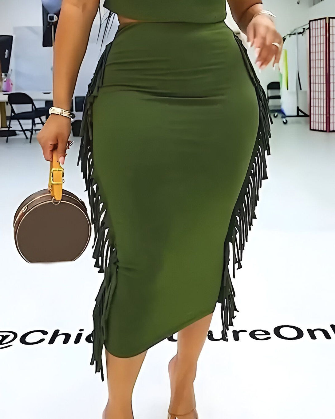 Une femme porte un ensemble deux pièces vert olive composé d'un crop top et d'une jupe fendue sur le côté avec des franges. Elle porte également des sandales à talons et un sac à main marron.