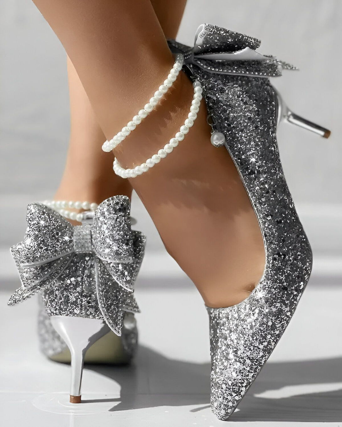 Une femme porte des escarpins argentés à talons hauts. Les escarpins sont décorés de nœuds en strass et de perles.