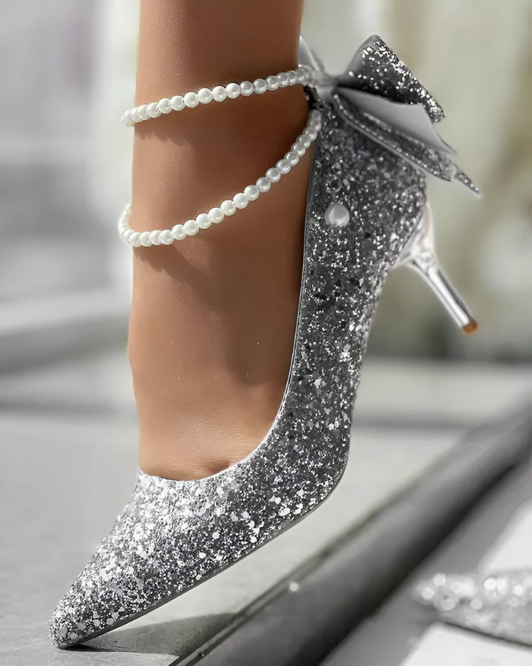 Une image d'une personne portant des escarpins argentés à talons hauts. Les escarpins sont recouverts de paillettes et ont un nœud en satin sur le devant. La personne porte également un bracelet de perles à la cheville.