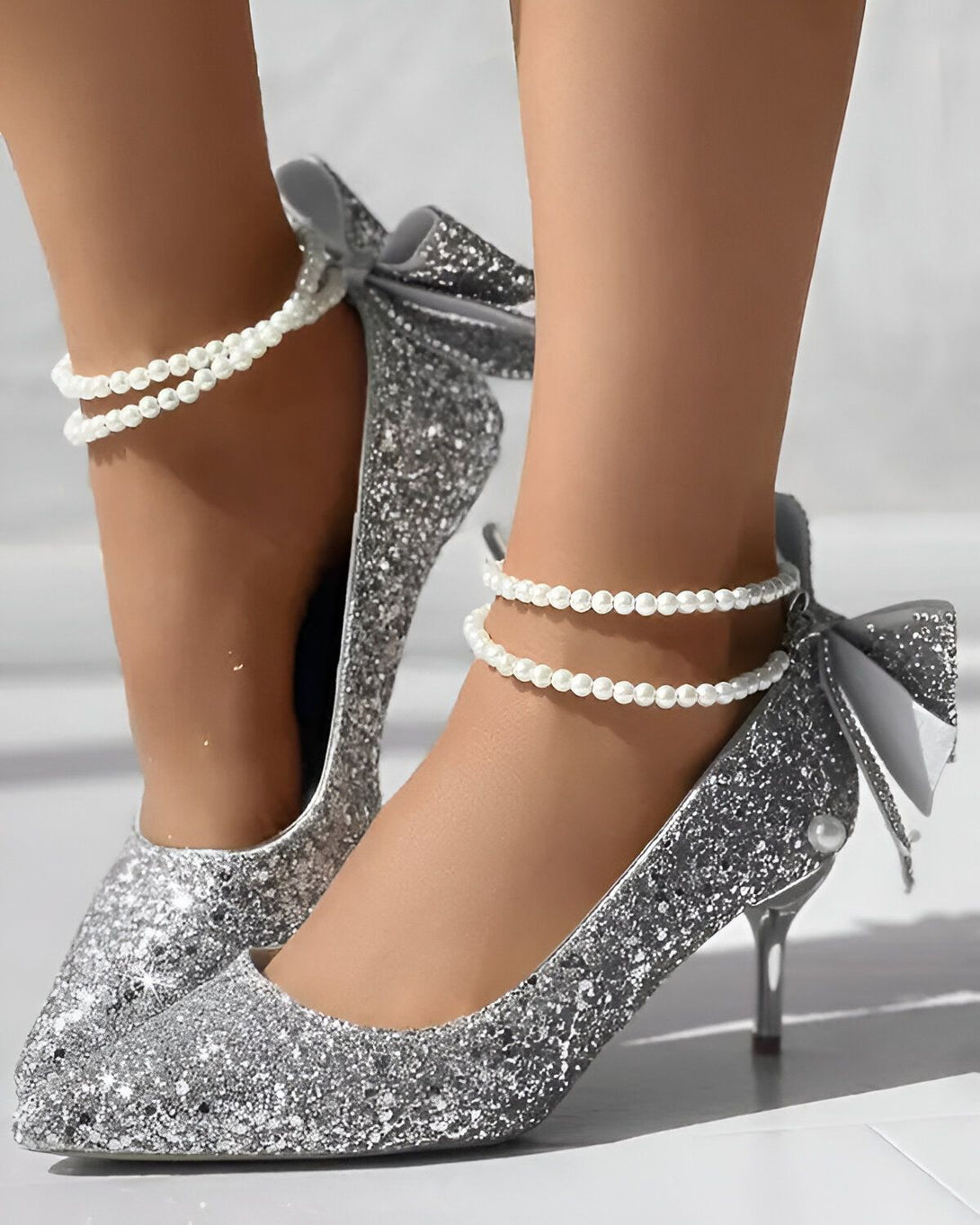 Une femme porte des escarpins argentés à talons hauts. Les escarpins sont recouverts de paillettes et ont un nœud en satin blanc sur le devant. La femme porte également des bracelets de perles autour des chevilles.
