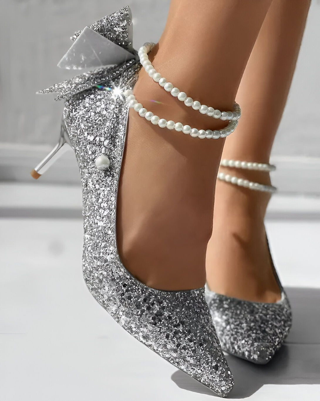 Une image d'une paire d'escarpins argentés à talons hauts avec un nœud en perles sur le devant. Les escarpins sont portés par une femme avec une robe blanche.