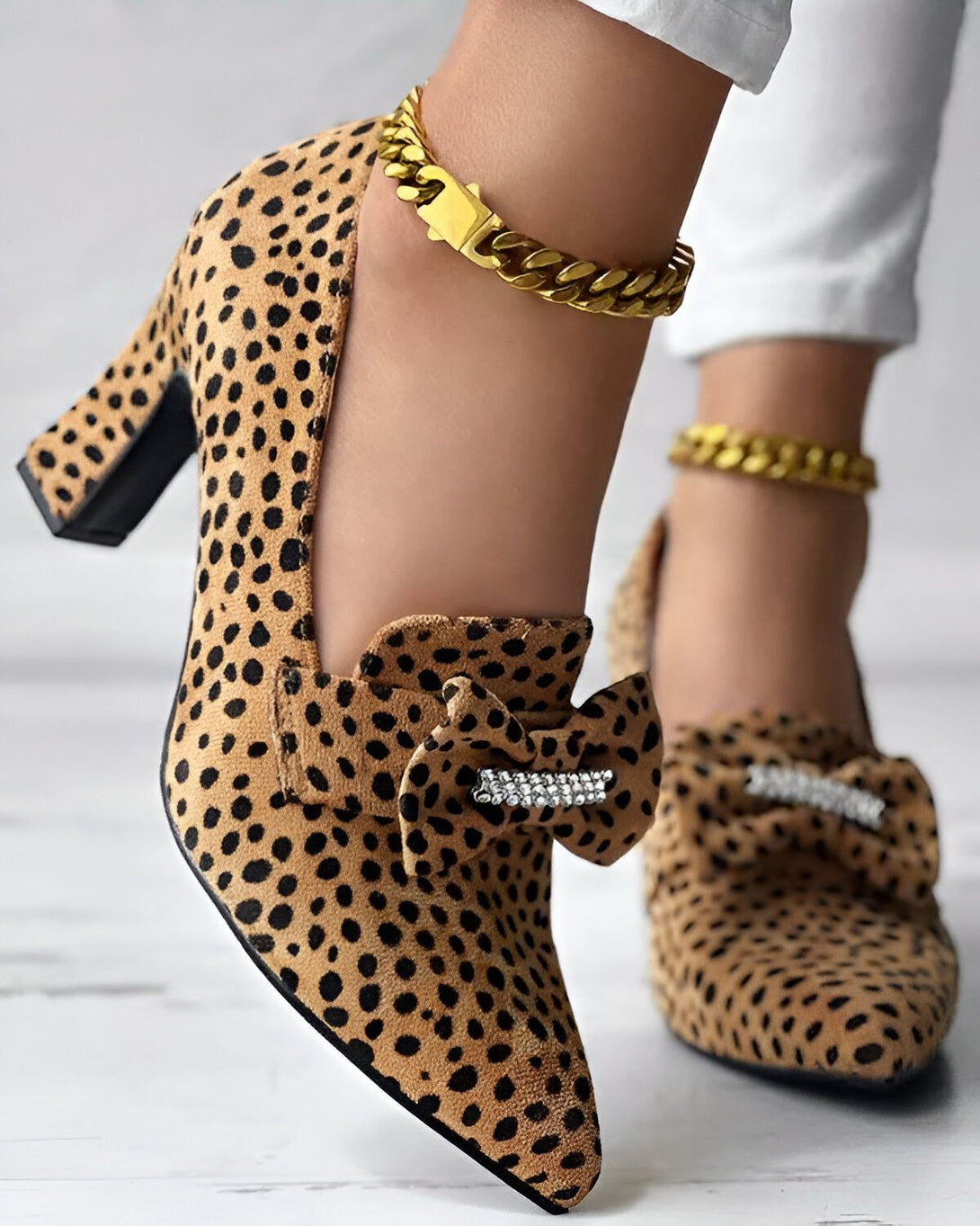 Une image d'une paire d'escarpins léopard à talons hauts avec un nœud en strass sur le devant. Les chaussures sont portées par une femme qui porte également un bracelet de cheville en chaîne dorée.