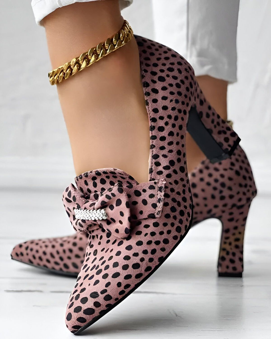 Une femme porte des escarpins roses à talons hauts. Les escarpins sont en daim et ont un motif à pois noirs. Il y a un nœud en strass sur le devant des escarpins. La femme porte également un pantalon blanc et un chemisier blanc. Elle a les cheveux longs et noirs. Elle est debout sur un sol en bois.