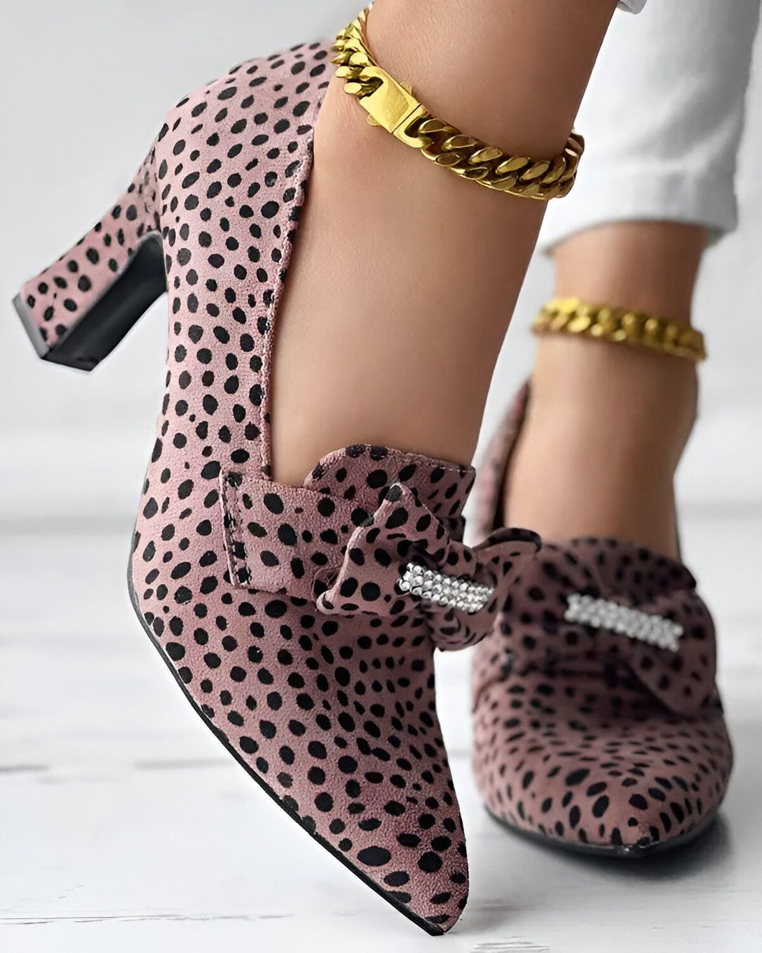 Une image de chaussures à talons roses avec des pois noirs et un nœud décoratif sur le devant. Les chaussures sont portées par une femme qui porte également un bracelet de cheville en or.