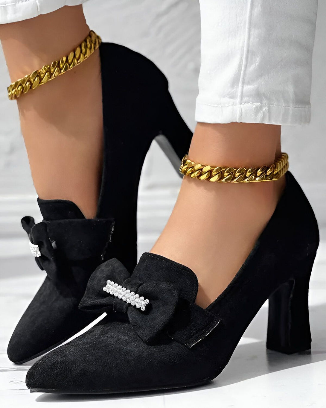 Une femme porte des escarpins noirs à talons hauts avec un nœud en strass sur le devant et une chaîne dorée autour de la cheville. Elle porte également un jean blanc.