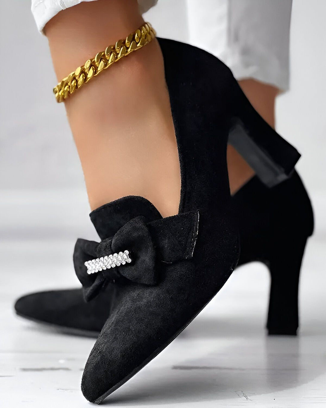 Une femme porte des escarpins noirs à talons hauts. Les escarpins sont en daim et ont un nœud en strass sur le devant. La femme porte également un bracelet de cheville en or.