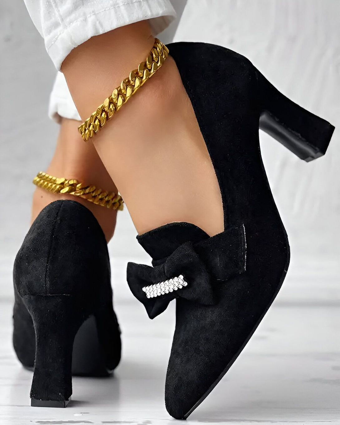 Une femme porte des escarpins noirs à talons hauts. Les escarpins sont en daim et ont un nœud en strass sur le devant. La femme porte également un pantalon blanc et des bijoux en or.