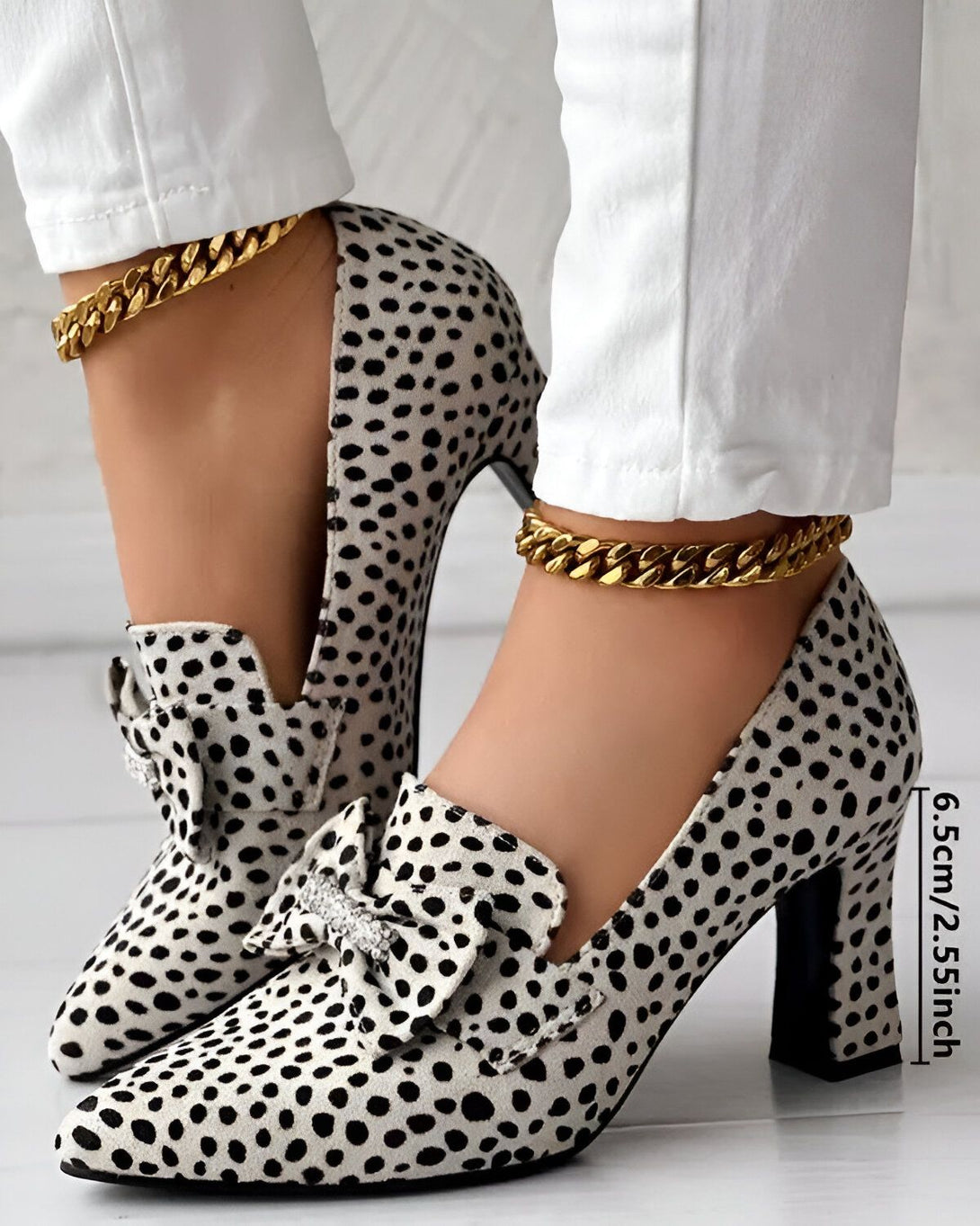 Une image de chaussures à talons hauts pour femmes. Les chaussures sont blanches à pois noirs et ont un nœud sur le devant. Le talon est épais et mesure 6,5 cm. Les chaussures sont portées par une femme qui porte un pantalon blanc et une chaîne dorée à la cheville.