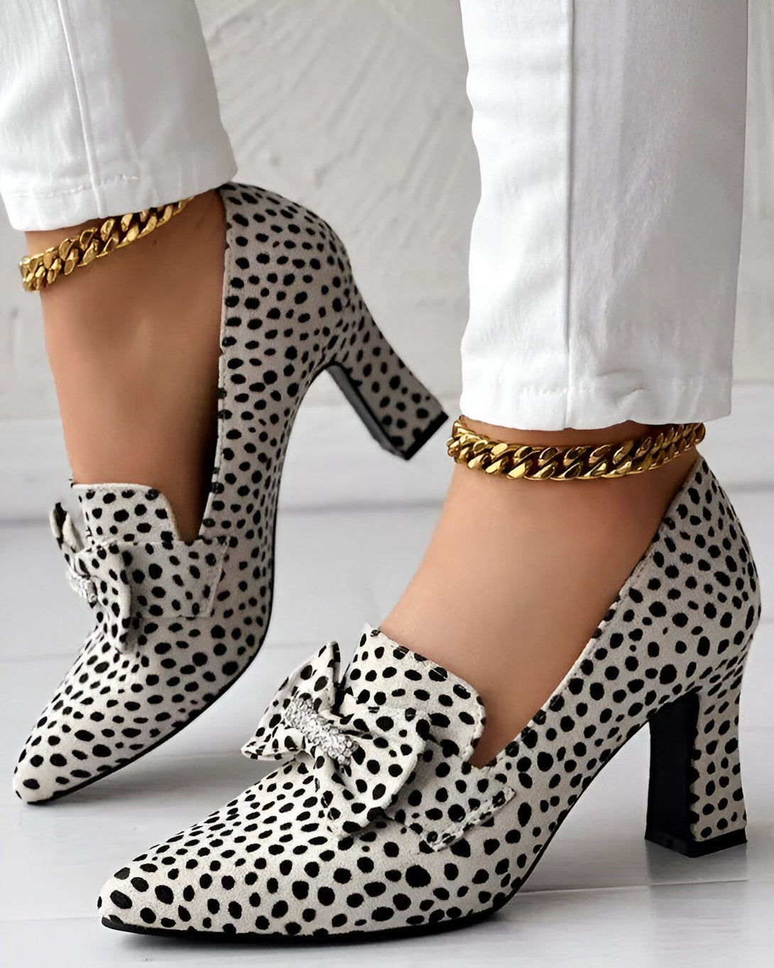 Une femme porte des escarpins noirs et blancs à talons hauts. Les escarpins sont en daim et ont un nœud sur le devant. La femme porte également un pantalon blanc et des bijoux dorés.