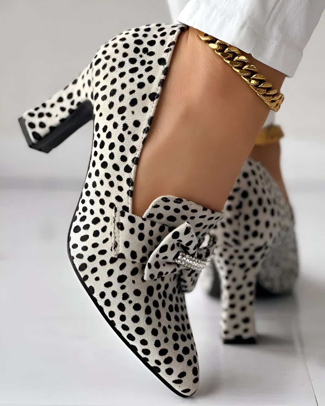 Une image d'une paire d'escarpins à talons hauts blancs à pois noirs. Les chaussures sont portées par une femme qui porte également un pantalon blanc et une cheville dorée.