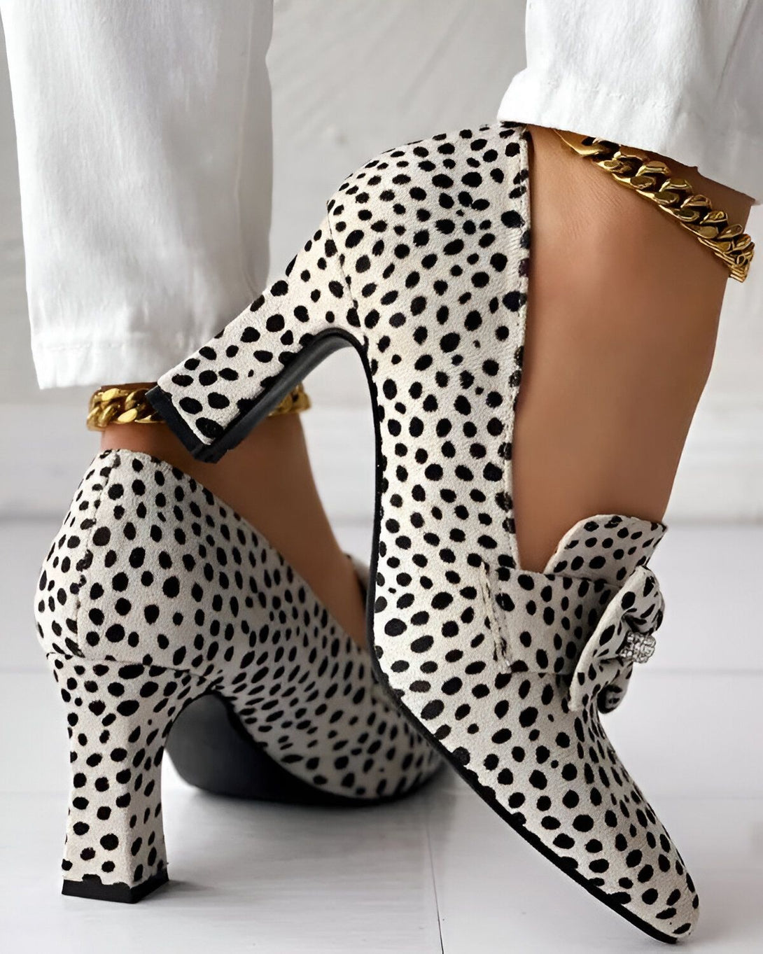 Une femme porte des escarpins blancs à talons hauts avec des pois noirs et une boucle en strass. Elle porte également un pantalon blanc et un bracelet de cheville doré.