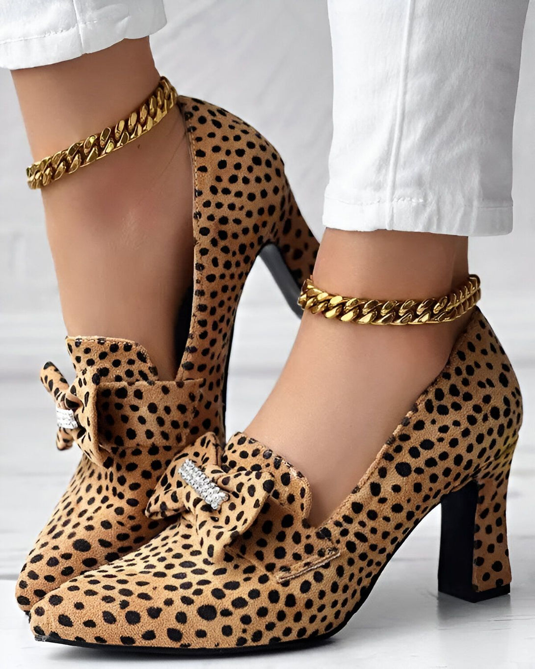 Une femme porte des escarpins léopard à talons hauts avec un nœud sur le devant et une chaîne dorée autour de la cheville. Elle porte également un jean blanc.