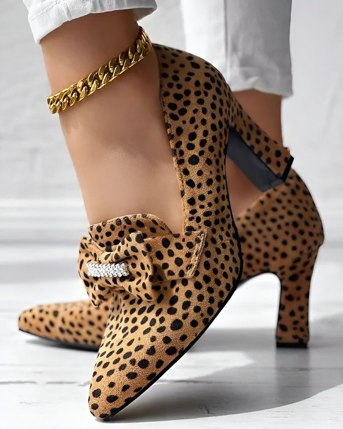 Une femme porte des escarpins léopard à talons hauts. Les chaussures ont un bout pointu et sont ornées d'un nœud en strass. La femme porte également un pantalon blanc et un bracelet de cheville doré.