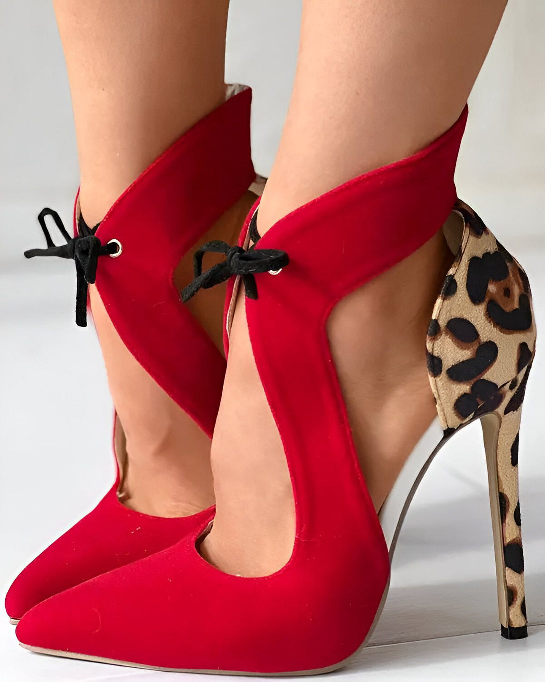 Une femme porte des escarpins rouges à talons hauts. Les escarpins sont ouverts sur le côté et ont un bout pointu. Le talon est fin et mesure environ 10 cm. La femme a les jambes croisées et porte une robe noire.