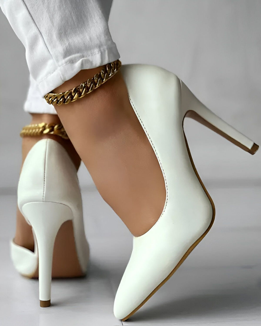 Une femme porte des escarpins blancs à talons hauts. Elle porte également une chaîne dorée autour de la cheville.